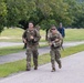 Two U.S. Army soldiers in MultiCam (OCP) camouflage uniforms are conducting a ruck march along a paved road on what appears to be a military installation. Both soldiers are carrying large rucksacks and appear to be in good spirits, with the soldier on the right smiling broadly. Additional personnel are visible in the background on the same road.