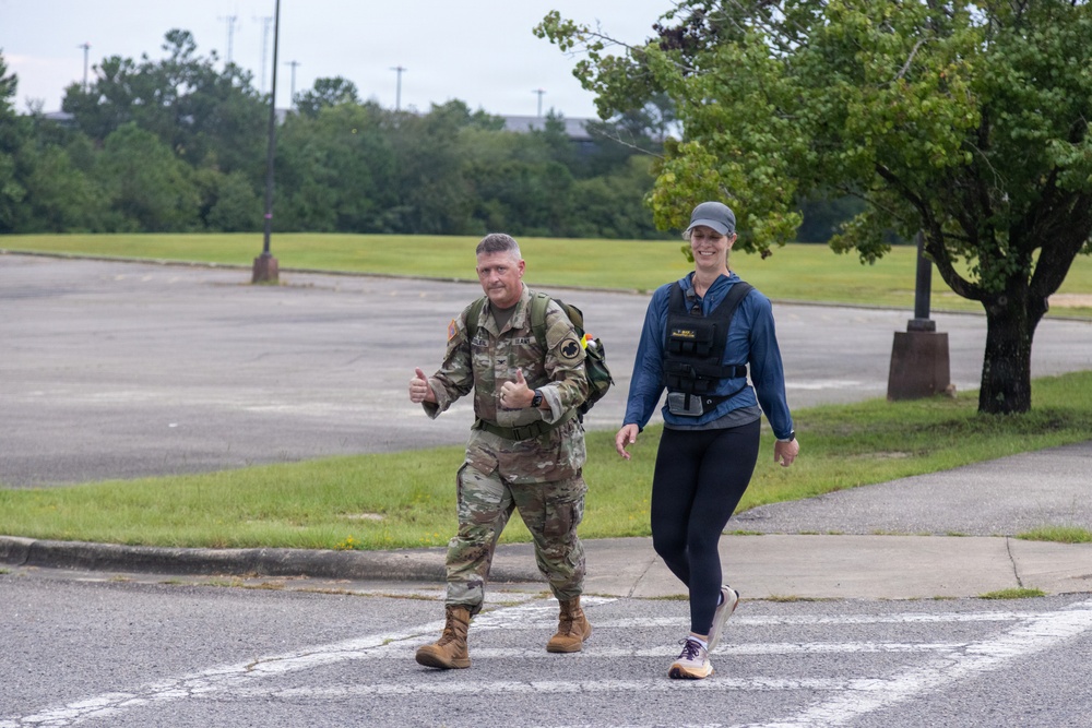 Two individuals are walking together across a crosswalk on what appears to be a military installation or base. One person is wearing full U.S. Army combat uniform (OCP/multicam) with a backpack and giving thumbs up, while the other is a civilian woman in athletic wear with a weighted vest. The setting suggests a ruck march or fitness event taking place outdoors.
