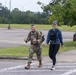 Two individuals are walking together across a crosswalk on what appears to be a military installation or base. One person is wearing full U.S. Army combat uniform (OCP/multicam) with a backpack and giving thumbs up, while the other is a civilian woman in athletic wear with a weighted vest. The setting suggests a ruck march or fitness event taking place outdoors.