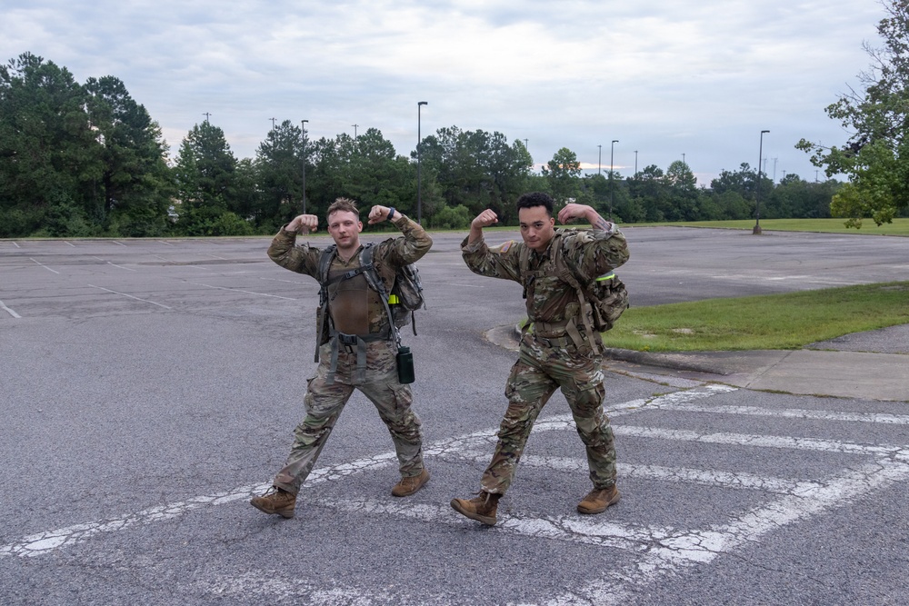 Two military personnel in full camouflage uniforms and tactical gear are posing in a parking lot, flexing their biceps for the camera. Both are wearing rucksacks and appear to be in good spirits, likely after completing a physical training exercise or ruck march. The setting appears to be a military installation or base facility.