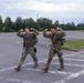 Two military personnel in full camouflage uniforms and tactical gear are posing in a parking lot, flexing their biceps for the camera. Both are wearing rucksacks and appear to be in good spirits, likely after completing a physical training exercise or ruck march. The setting appears to be a military installation or base facility.