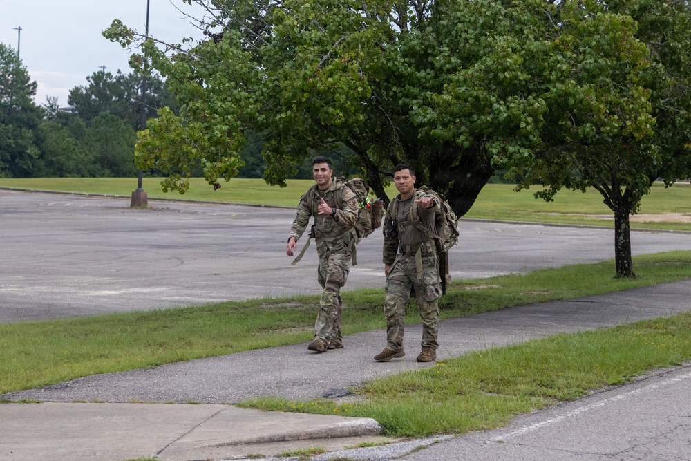 Two military personnel in camouflage uniforms are walking along a paved path on what appears to be a military installation or base. Both soldiers are carrying large rucksacks/backpacks and appear to be in good spirits, with one smiling at the camera. The setting includes an open parking lot, green grass, and mature trees under an overcast sky.