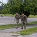Two military personnel in camouflage uniforms are walking along a paved path on what appears to be a military installation or base. Both soldiers are carrying large rucksacks/backpacks and appear to be in good spirits, with one smiling at the camera. The setting includes an open parking lot, green grass, and mature trees under an overcast sky.