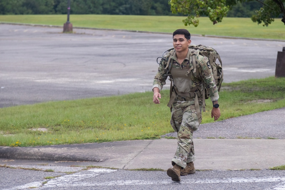 A young soldier in US Army OCP (Operational Camouflage Pattern) uniform is walking briskly along a sidewalk while carrying a large military rucksack. The individual appears to be participating in a ruck march or physical training exercise, smiling as they move. The setting appears to be a military installation with open fields and a parking area visible in the background.