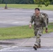 A young soldier in US Army OCP (Operational Camouflage Pattern) uniform is walking briskly along a sidewalk while carrying a large military rucksack. The individual appears to be participating in a ruck march or physical training exercise, smiling as they move. The setting appears to be a military installation with open fields and a parking area visible in the background.