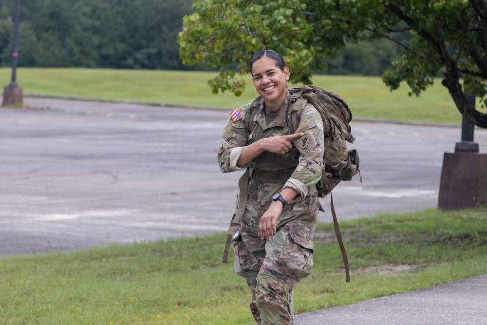 A female U.S. Army soldier in Operational Camouflage Pattern (OCP) uniform is smiling broadly while carrying a large military rucksack. She is pointing toward her shoulder patch and appears to be in motion, likely during a ruck march or physical training exercise. The setting appears to be a military installation with open fields and trees in the background.