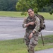 A female U.S. Army soldier in Operational Camouflage Pattern (OCP) uniform is smiling broadly while carrying a large military rucksack. She is pointing toward her shoulder patch and appears to be in motion, likely during a ruck march or physical training exercise. The setting appears to be a military installation with open fields and trees in the background.