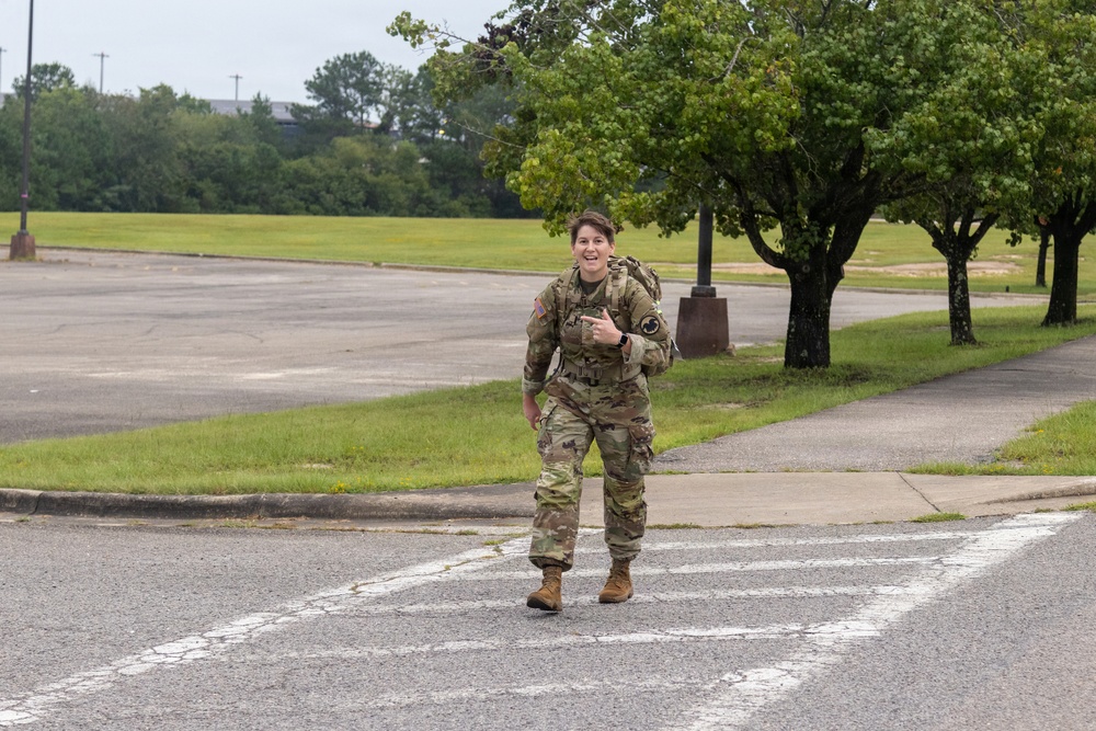 A female soldier in U.S. Army MultiCam (OCP) camouflage uniform is walking across a road on what appears to be a military installation. She is smiling and making a pointing gesture toward the camera while carrying a rucksack on her back. The setting includes a parking lot, grassy areas, and trees under an overcast sky.