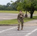 A female soldier in U.S. Army MultiCam (OCP) camouflage uniform is walking across a road on what appears to be a military installation. She is smiling and making a pointing gesture toward the camera while carrying a rucksack on her back. The setting includes a parking lot, grassy areas, and trees under an overcast sky.
