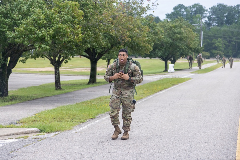 A soldier in full camouflage uniform and tactical backpack is rucking (marching with a loaded pack) along a paved road on what appears to be a military installation. The soldier in the foreground appears to be exerting significant effort, with a strained facial expression. Several other soldiers in similar gear are visible in the background along the same road.