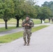 A soldier in full camouflage uniform and tactical backpack is rucking (marching with a loaded pack) along a paved road on what appears to be a military installation. The soldier in the foreground appears to be exerting significant effort, with a strained facial expression. Several other soldiers in similar gear are visible in the background along the same road.