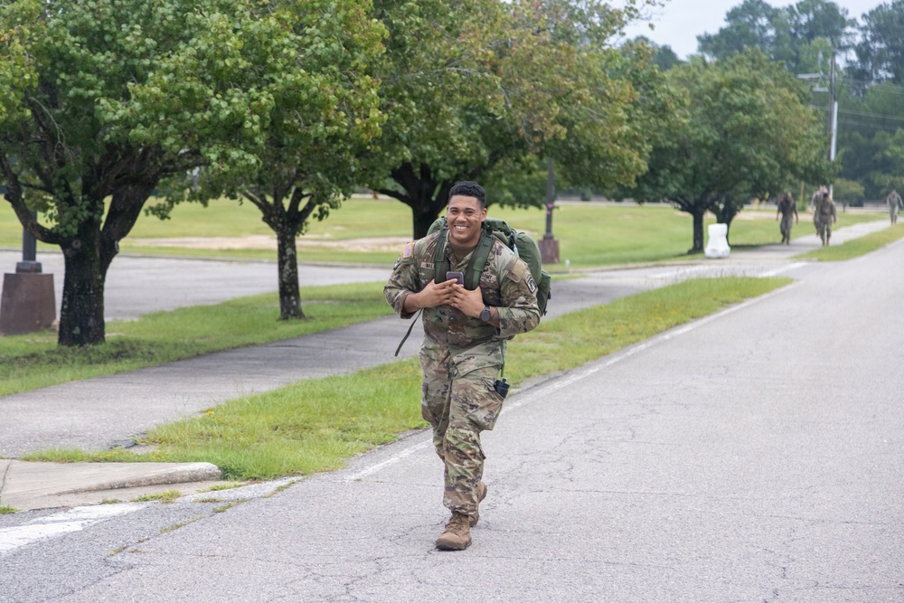 A smiling soldier in full camouflage uniform and tactical gear is walking or jogging along a road on what appears to be a military installation. He is carrying a large green rucksack on his back and holding a smartphone in his hands. Additional soldiers are visible in the background along the same road, suggesting a group ruck march or physical training event.