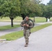 A smiling soldier in full camouflage uniform and tactical gear is walking or jogging along a road on what appears to be a military installation. He is carrying a large green rucksack on his back and holding a smartphone in his hands. Additional soldiers are visible in the background along the same road, suggesting a group ruck march or physical training event.
