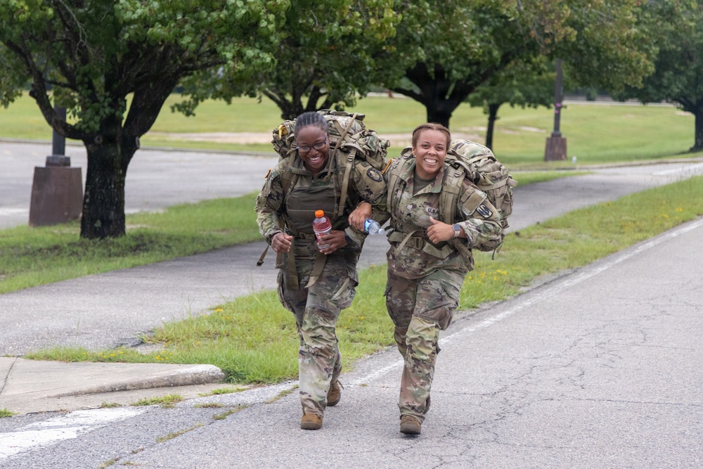 Two female soldiers in U.S. Army OCP (Operational Camouflage Pattern) uniforms are running along a paved road while carrying large rucksacks on their backs. Both are smiling broadly, suggesting high morale during what appears to be a ruck march or physical training exercise. The setting appears to be a military installation with manicured grounds and trees in the background.