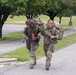 Two female soldiers in U.S. Army OCP (Operational Camouflage Pattern) uniforms are running along a paved road while carrying large rucksacks on their backs. Both are smiling broadly, suggesting high morale during what appears to be a ruck march or physical training exercise. The setting appears to be a military installation with manicured grounds and trees in the background.