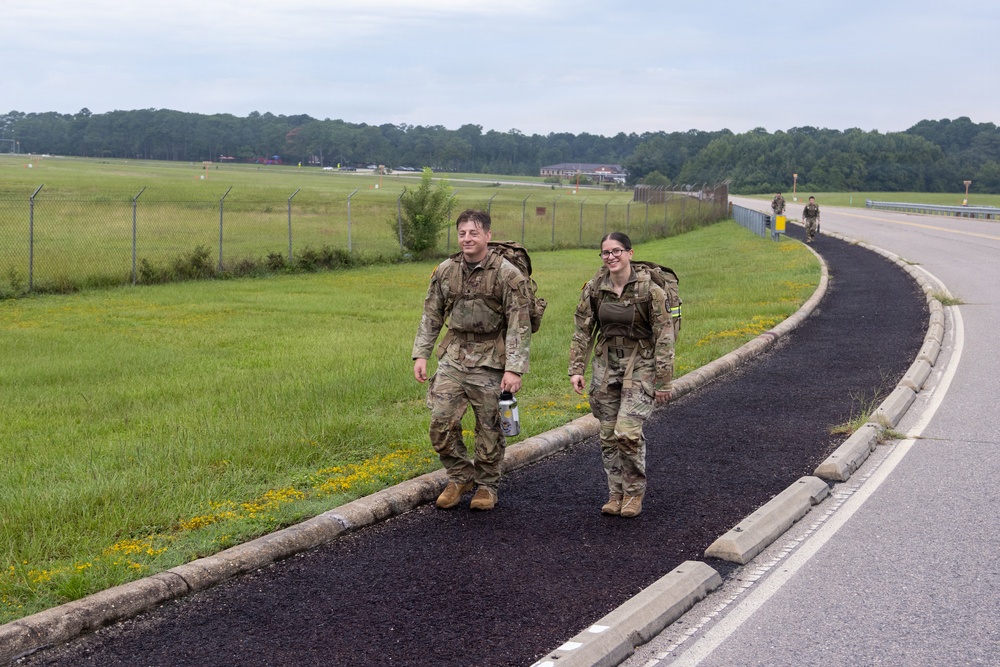 Two military personnel in camouflage uniforms and rucksacks are walking along a paved path beside a road on what appears to be a military installation. Both are smiling and appear to be participating in a ruck march or similar physical training exercise. At least one additional service member is visible in the background further down the path.