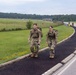 Two military personnel in camouflage uniforms and rucksacks are walking along a paved path beside a road on what appears to be a military installation. Both are smiling and appear to be participating in a ruck march or similar physical training exercise. At least one additional service member is visible in the background further down the path.