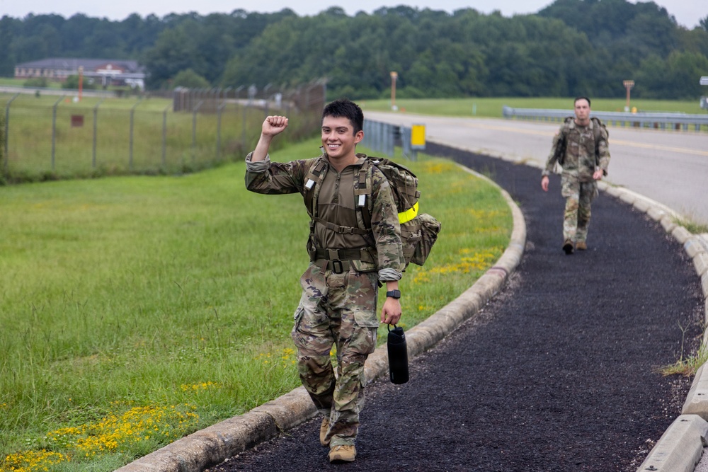 Two military personnel in camouflage uniforms are walking along a paved path on what appears to be a military installation or airfield. The soldier in the foreground is smiling and raising a fist in a celebratory gesture, carrying a water bottle and wearing a tactical vest with a yellow reflective band. The background shows a grassy airfield area, fencing, a road, and dense tree line.