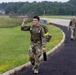 Two military personnel in camouflage uniforms are walking along a paved path on what appears to be a military installation or airfield. The soldier in the foreground is smiling and raising a fist in a celebratory gesture, carrying a water bottle and wearing a tactical vest with a yellow reflective band. The background shows a grassy airfield area, fencing, a road, and dense tree line.