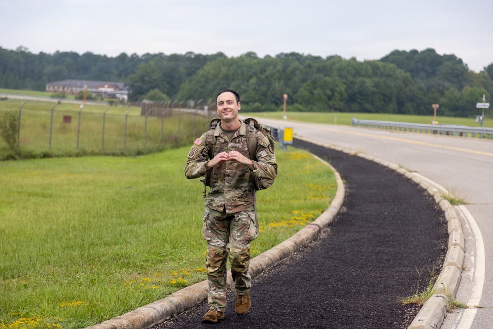 A U.S. Army soldier in full Operational Camouflage Pattern (OCP) uniform walks along a paved path or road on what appears to be a military installation. The soldier is smiling and making a heart shape with his hands toward the camera while carrying a rucksack. The setting includes green grass, a perimeter fence, trees, and military-style buildings in the background.