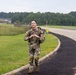 A U.S. Army soldier in full Operational Camouflage Pattern (OCP) uniform walks along a paved path or road on what appears to be a military installation. The soldier is smiling and making a heart shape with his hands toward the camera while carrying a rucksack. The setting includes green grass, a perimeter fence, trees, and military-style buildings in the background.