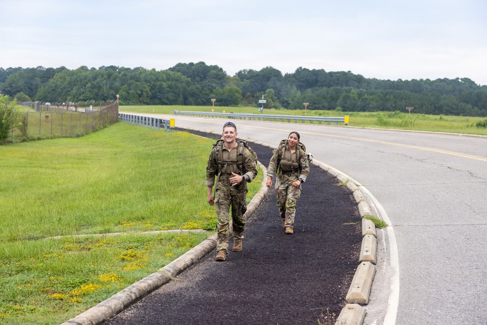 Two military personnel in full combat uniforms and rucksacks are walking along a paved path beside a road, both smiling. The setting appears to be a military installation or base, identifiable by the fenced perimeter, open fields, and runway-style infrastructure in the background. The image captures what appears to be a ruck march or physical training exercise.