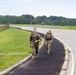 Two military personnel in full combat uniforms and rucksacks are walking along a paved path beside a road, both smiling. The setting appears to be a military installation or base, identifiable by the fenced perimeter, open fields, and runway-style infrastructure in the background. The image captures what appears to be a ruck march or physical training exercise.