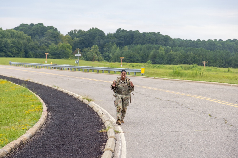 A female soldier in full camouflage uniform and tactical gear is walking along a road on what appears to be a military installation or training area. She is smiling and making peace signs with both hands, suggesting she is completing or near the end of a ruck march or physical training exercise. The setting is a rural, green landscape with trees in the background under an overcast sky.
