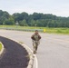 A female soldier in full camouflage uniform and tactical gear is walking along a road on what appears to be a military installation or training area. She is smiling and making peace signs with both hands, suggesting she is completing or near the end of a ruck march or physical training exercise. The setting is a rural, green landscape with trees in the background under an overcast sky.