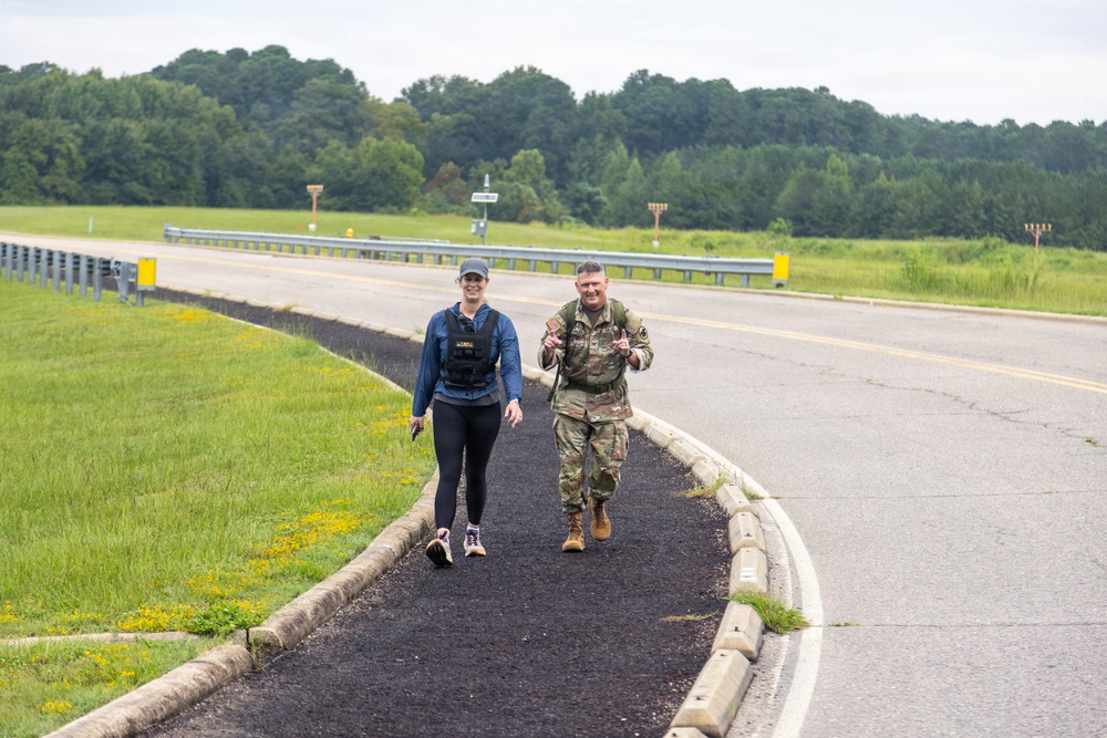 Two individuals are walking along a paved path beside a road in what appears to be a military installation or base. One person is wearing civilian athletic clothing with a weighted vest, while the other is dressed in U.S. Army camouflage (OCP) uniform with a rucksack or tactical vest. The setting features open green fields, a guardrail, and a tree line in the background under an overcast sky.