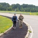 Two individuals are walking along a paved path beside a road in what appears to be a military installation or base. One person is wearing civilian athletic clothing with a weighted vest, while the other is dressed in U.S. Army camouflage (OCP) uniform with a rucksack or tactical vest. The setting features open green fields, a guardrail, and a tree line in the background under an overcast sky.