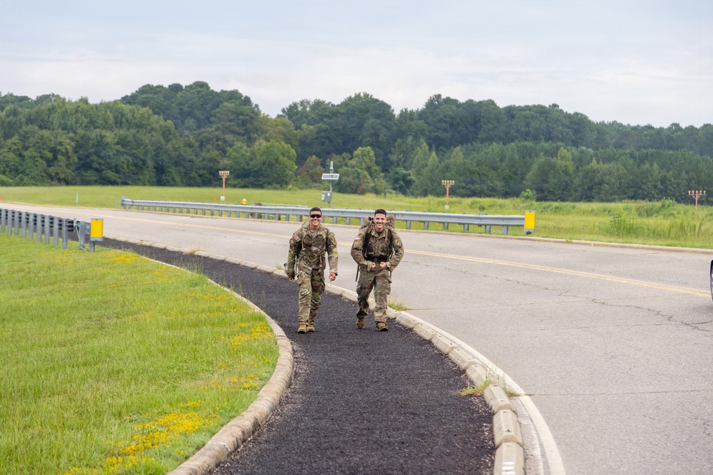 Two military personnel in camouflage uniforms are walking/jogging along a paved pathway beside a road, both smiling and appearing to be in good spirits. At least one individual is carrying a rucksack, suggesting they may be completing a ruck march or physical training exercise. The setting appears to be a military installation or base, with open grassy areas, guardrails, and runway-style lighting structures visible in the background.