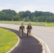 Two military personnel in camouflage uniforms are walking/jogging along a paved pathway beside a road, both smiling and appearing to be in good spirits. At least one individual is carrying a rucksack, suggesting they may be completing a ruck march or physical training exercise. The setting appears to be a military installation or base, with open grassy areas, guardrails, and runway-style lighting structures visible in the background.