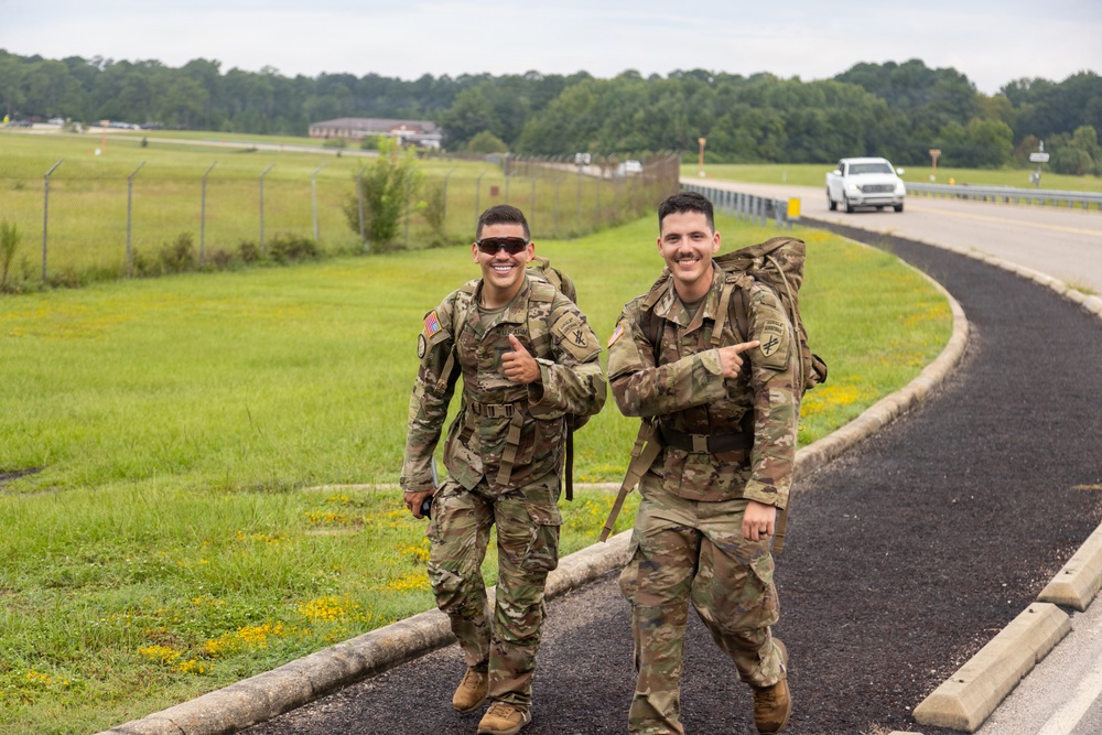 Two U.S. Army soldiers in OCP (Operational Camouflage Pattern) uniforms are walking along a paved path on what appears to be a military installation, both carrying large rucksacks and smiling at the camera. One soldier is giving a thumbs-up while the other points toward his companion. The setting appears to be a ruck march or physical training event on a military base.