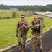 Two U.S. Army soldiers in OCP (Operational Camouflage Pattern) uniforms are walking along a paved path on what appears to be a military installation, both carrying large rucksacks and smiling at the camera. One soldier is giving a thumbs-up while the other points toward his companion. The setting appears to be a ruck march or physical training event on a military base.