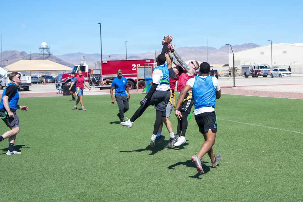 A group of players are engaged in a flag football game on an artificial turf field, with multiple players jumping to contest a pass near the center of the action. The game appears to be an informal or recreational league event, with players wearing blue and red colored pinnies/vests to distinguish teams. The setting appears to be a military base or similar facility, evidenced by fire trucks, hangars, and desert mountain terrain visible in the background.