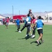A group of players are engaged in a flag football game on an artificial turf field, with multiple players jumping to contest a pass near the center of the action. The game appears to be an informal or recreational league event, with players wearing blue and red colored pinnies/vests to distinguish teams. The setting appears to be a military base or similar facility, evidenced by fire trucks, hangars, and desert mountain terrain visible in the background.