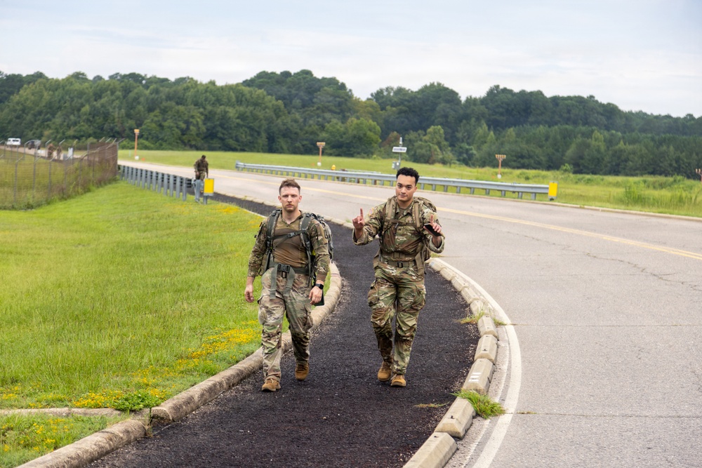 Two soldiers in camouflage uniforms and tactical gear are walking along a paved path beside a road, with one making a peace/victory hand gesture toward the camera. A third soldier is visible in the background further down the road. The setting appears to be a military installation or training area, surrounded by green grass and wooded treelines.