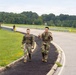Two soldiers in camouflage uniforms and tactical gear are walking along a paved path beside a road, with one making a peace/victory hand gesture toward the camera. A third soldier is visible in the background further down the road. The setting appears to be a military installation or training area, surrounded by green grass and wooded treelines.