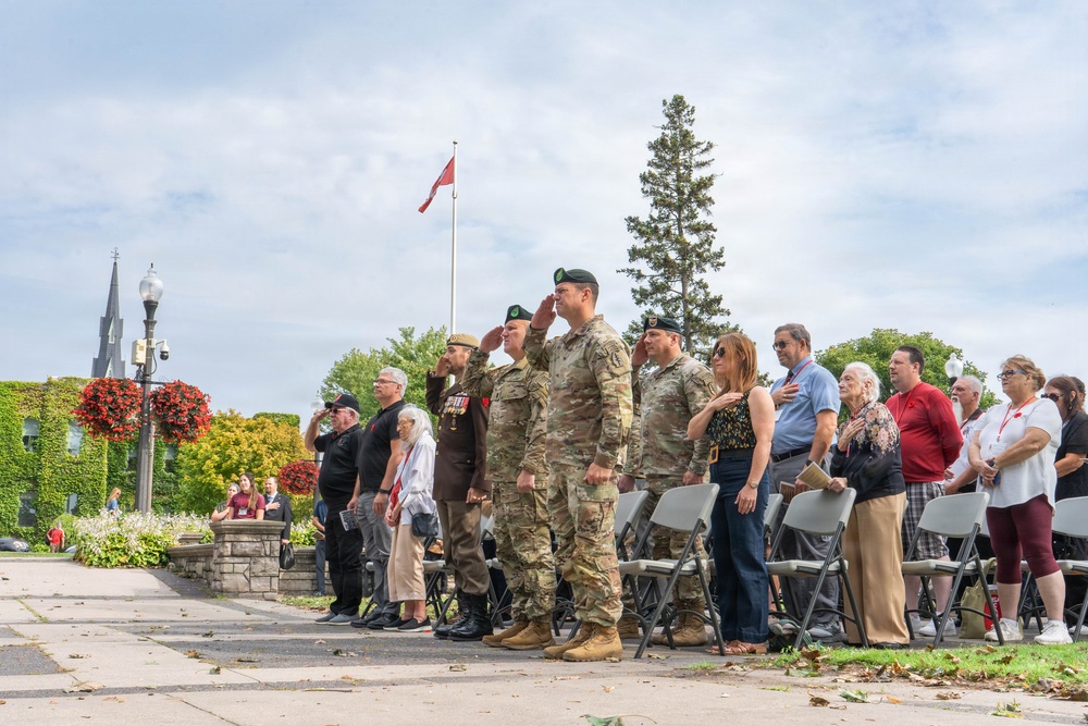 A military memorial or remembrance ceremony is taking place outdoors in what appears to be a Canadian town square or park. Several military personnel in camouflage uniforms and berets are saluting, while civilians stand respectfully, some with hands over their hearts. A Canadian flag flies at half-mast in the background, suggesting a solemn commemorative occasion.
