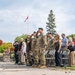 A military memorial or remembrance ceremony is taking place outdoors in what appears to be a Canadian town square or park. Several military personnel in camouflage uniforms and berets are saluting, while civilians stand respectfully, some with hands over their hearts. A Canadian flag flies at half-mast in the background, suggesting a solemn commemorative occasion.