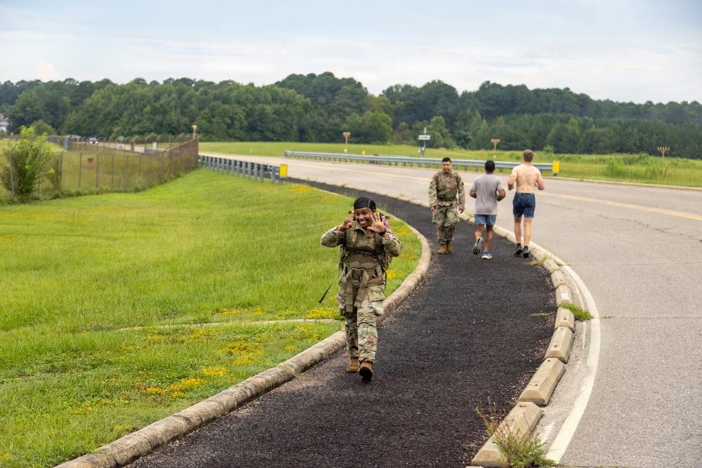 A group of military personnel and civilians are exercising on a paved path alongside a road, likely on a military base. A soldier in full camouflage uniform and tactical gear is walking toward the camera, smiling and waving. Behind her, two soldiers in uniform and two civilians in casual athletic wear are running away from the camera.