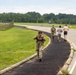A group of military personnel and civilians are exercising on a paved path alongside a road, likely on a military base. A soldier in full camouflage uniform and tactical gear is walking toward the camera, smiling and waving. Behind her, two soldiers in uniform and two civilians in casual athletic wear are running away from the camera.
