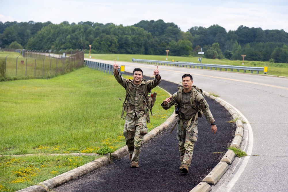 Two soldiers in U.S. Army MultiCam/OCP camouflage uniforms are walking along a paved path beside a road on what appears to be a military installation. Both are wearing full tactical gear including rucksacks, suggesting they are completing a ruck march or similar physical training event. The soldier in front is smiling and raising both index fingers upward in a celebratory gesture.