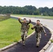 Two soldiers in U.S. Army MultiCam/OCP camouflage uniforms are walking along a paved path beside a road on what appears to be a military installation. Both are wearing full tactical gear including rucksacks, suggesting they are completing a ruck march or similar physical training event. The soldier in front is smiling and raising both index fingers upward in a celebratory gesture.