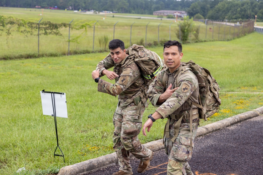 Two U.S. Army soldiers in OCP (Operational Camouflage Pattern) uniforms are conducting what appears to be a ruck march or physical fitness event on a paved path near a grassy field. Both soldiers are carrying large military rucksacks and appear to be in good spirits, with one pointing toward the camera. The setting appears to be a military installation, visible by the perimeter fencing and maintained grounds in the background.