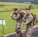 Two U.S. Army soldiers in OCP (Operational Camouflage Pattern) uniforms are conducting what appears to be a ruck march or physical fitness event on a paved path near a grassy field. Both soldiers are carrying large military rucksacks and appear to be in good spirits, with one pointing toward the camera. The setting appears to be a military installation, visible by the perimeter fencing and maintained grounds in the background.