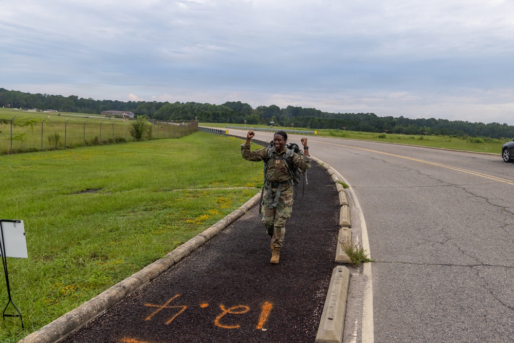 A soldier in military camouflage uniform and full rucksack is walking along a paved path beside a road, raising both fists in a celebratory gesture. The setting appears to be a military installation with open fields, a fence line, and tree lines in the background. Orange painted markings on the path read '18.1' (possibly mirrored/reversed), suggesting a distance marker for a ruck march event.