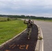 A soldier in military camouflage uniform and full rucksack is walking along a paved path beside a road, raising both fists in a celebratory gesture. The setting appears to be a military installation with open fields, a fence line, and tree lines in the background. Orange painted markings on the path read '18.1' (possibly mirrored/reversed), suggesting a distance marker for a ruck march event.