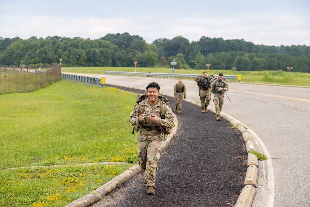 A group of military personnel in camouflage uniforms are conducting a ruck march along a paved path beside a road on what appears to be a military installation. The lead soldier, a smiling young man of apparent Asian descent, is carrying a large rucksack and appears in good spirits. Three additional soldiers follow behind him in a loose formation.